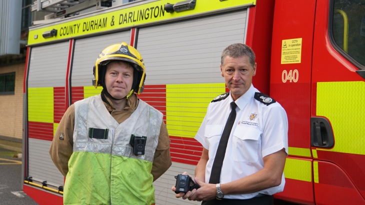 Chief Fire Officer Steve Helps holding a Body Worn Video Camera, alongside a Firefighter, stood In front of a fire truck