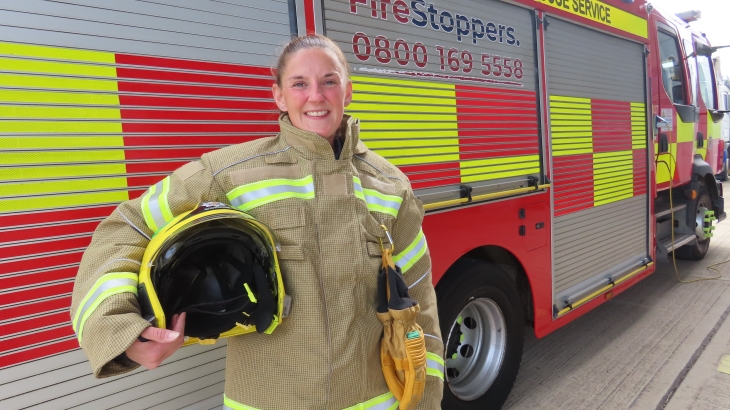 On-call firefighter Tracey Laws in fire kit holding helmet and standing in front of a g fire engine. 