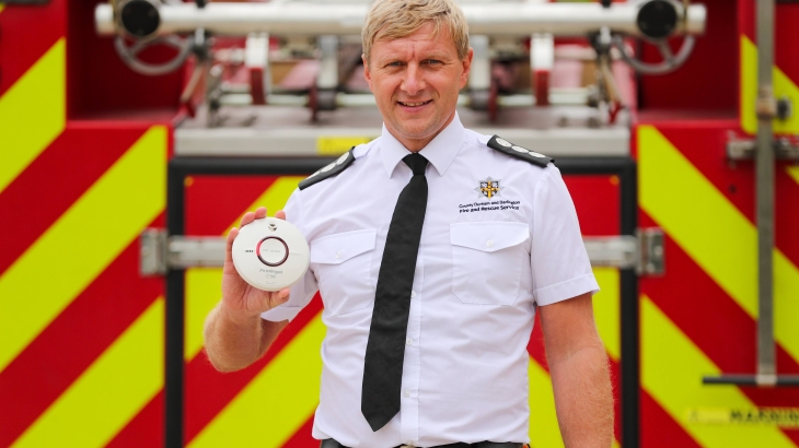 Station Manager Kevin Bell holding a smoke alarm and standing in front of a fire engine.