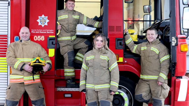 Four firefighters wearing fire kit and standing by a fire engine. 
