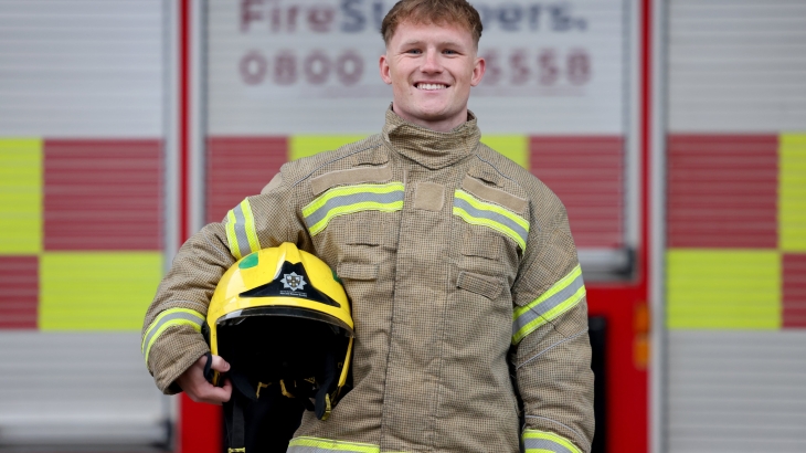 Crook on-call firefighter Kory Whitfield wearing fire kit and holding helmet under arm with fire engine in the background.