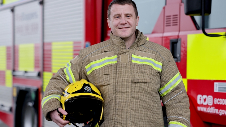 On-call firefighter Mark Thexton wearing fire kit with helmet under arm and fire engine in background. 