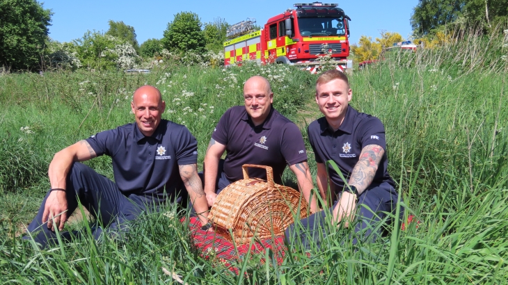 Three firefighters sitting on grass with a picnic and fire engine in the background. 