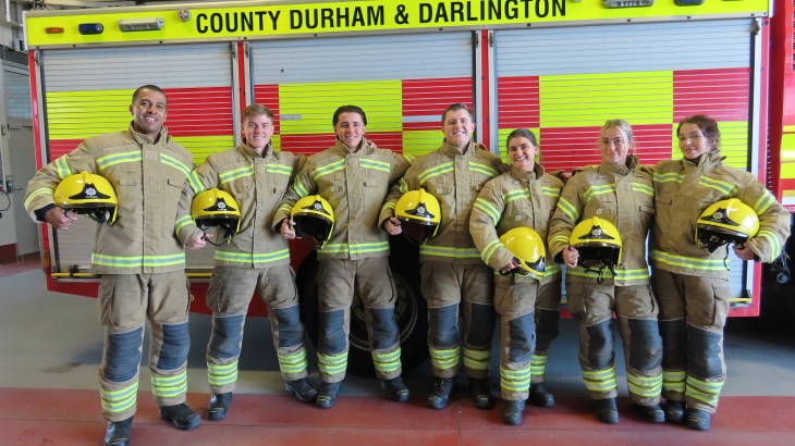 Seven firefighters from Cohort 8 wearing fire kit with helmets under arms and engine behind. 