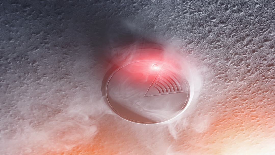 Smoke detector on a ceiling with a red light flashing, surrounded by a light smoke cloud.