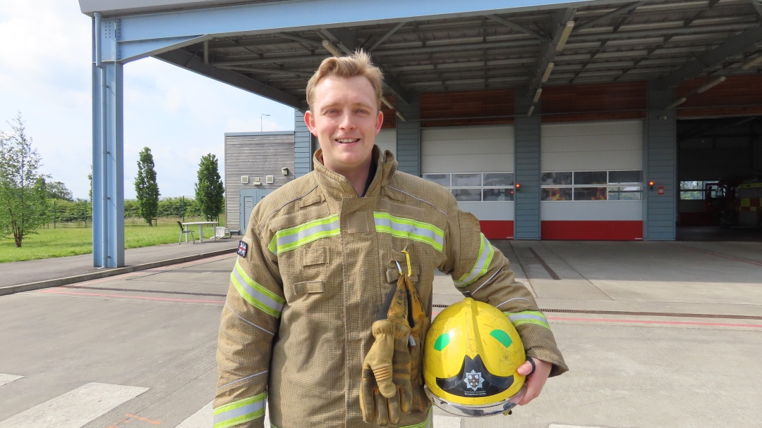 On-call firefighter Connor Turner wearing fire kit and holding helmet under his arm as he stands outside in the yard at Durham Fire Station. 