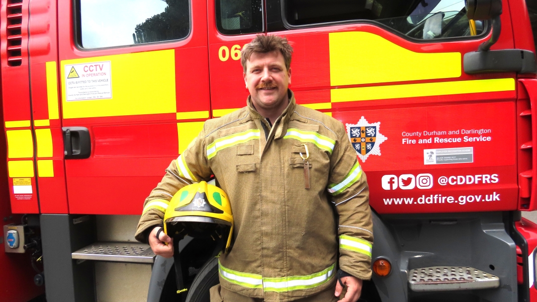 On-call firefighter Connor Mallinson wearing fire kit and holding his helmet in front of a fire engine. 