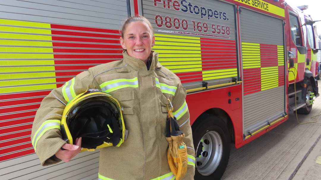 On-call firefighter Tracey Laws in fire kit holding helmet and standing in front of a g fire engine. 
