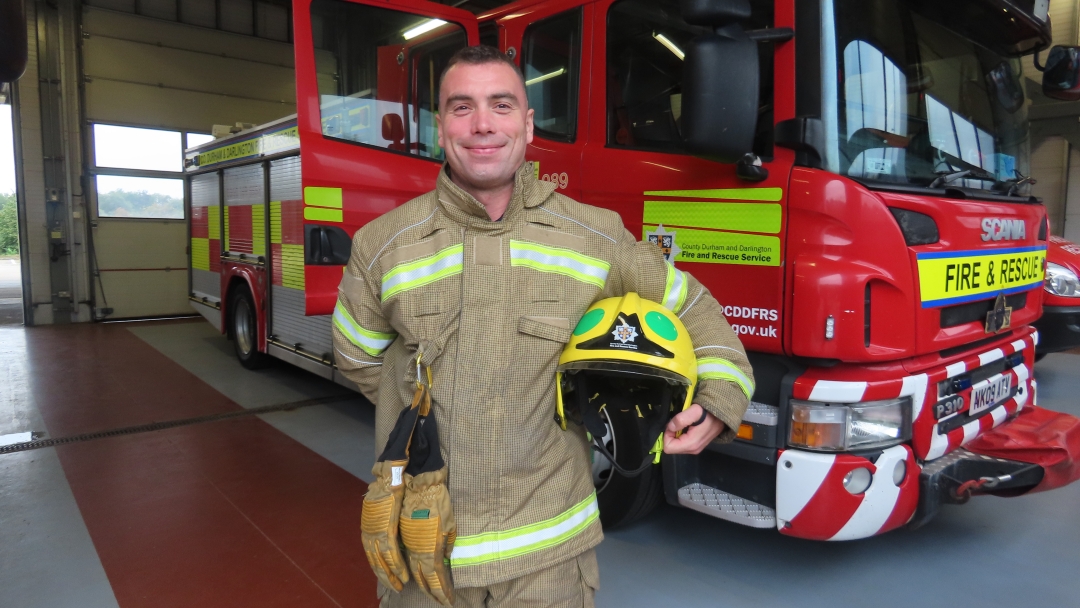 On-call firefighter Dan Hale wearing fire kit and holding helmet standing in front of a fire engine. 