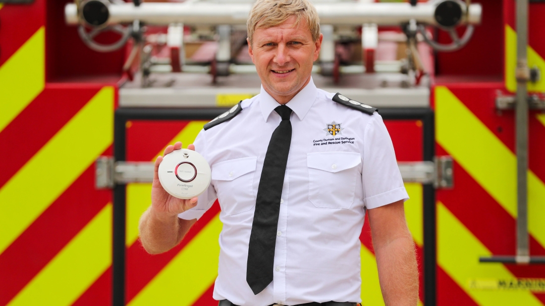 Station Manager Kevin Bell holding a smoke alarm and standing in front of a fire engine.