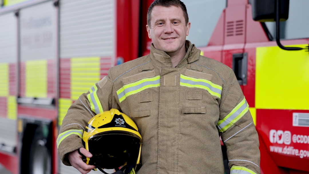 On-call firefighter Mark Thexton wearing fire kit with helmet under arm and fire engine in background. 